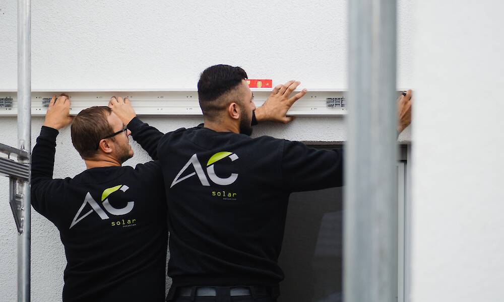 Zwei Arbeiter von "AC Solar Solution" montieren eine Schiene an einer Hauswand und verwenden dabei eine Wasserwaage.