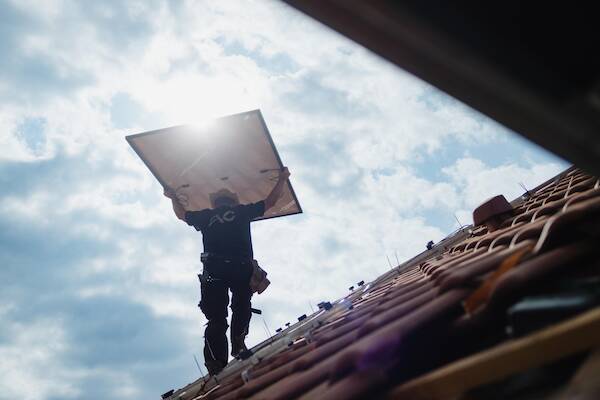 Professionelle Installation einer Photovoltaikanlage auf einem Ziegeldach: Qualifizierter Solarteur hebt ein Solarmodul zur Montage.